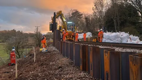 In failing light, a mechanical digger rests on stones on a rail line driving sheet metal piles into an embankment. Uniformed workers, other machinery and large sacks of ballast surround it. There are trees and a field either side of the railway.