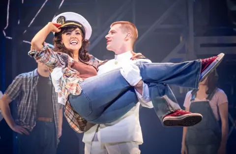 A young woman wearing jeans, a shirt and a sailor's cap is held in the arms of a man in a white sailor's uniform during a production of An Officer and a Gentleman at Bristol Hippodrome