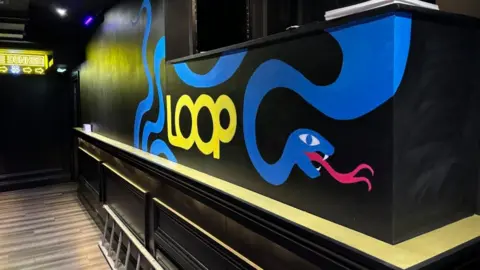 Charlie Stubbs / BBC There is a black wall and a brown wooden floor. On the wall, there is the artwork of a blue snake with yellow lettering saying 'Loop'. There is also a yellow topped ridge attached to the wall.