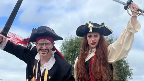 A man, on the left, and a woman, on the right, both looking at the camera and wearing pirate costumes 
