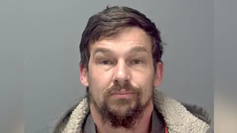 Mark Ling looks directly at the camera while being photographed for a custody image. He is wearing a grey fleece-lined hoodie and grey shirt. He has a dark beard and dark hair.  