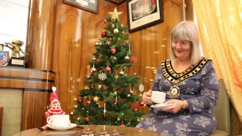 Swindon Borough Council Small elf with a cup of tea next to an older woman with mayoral chains holding a cup of tea. A Christmas tree is between them