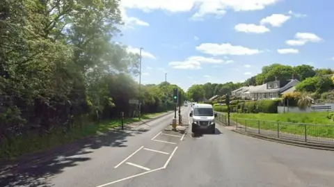 A Google street view screenshot of the A694 Lockhaugh Road, Rowlands Gill, Gateshead. There are trees either side of the road and the sky is blue and cloudy. 