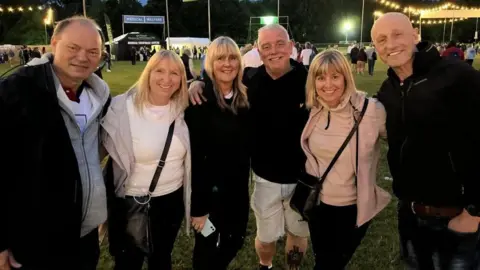 Mark Reynolds and five others smiling as they pose for a picture in a park. Other music fans are milling around behind them