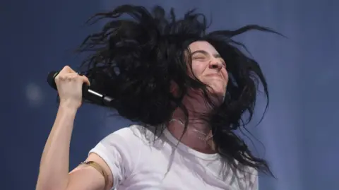 A close up of singer-songwriter Lorde. She has dark hair, is wearing a white T-shirt and is holding a microphone in one hand.