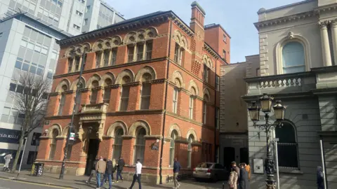 Outside view of 28 Bedford Street. It is a three-storey with attic Venetian Gothic redbrick and sandstone former linen store, dated 1867. People are walking on the street outside.