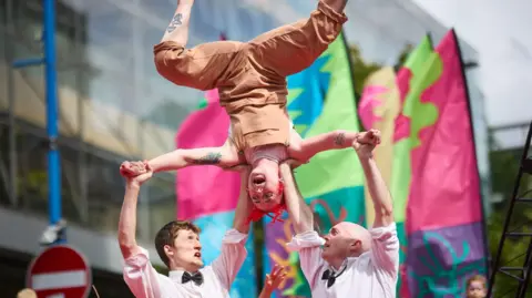 Mark Waugh Two men dressed in white shirts and dickie bow ties hold a woman up in the air who is upside down wearing beige dungarees. It is part of a street art performance. 
