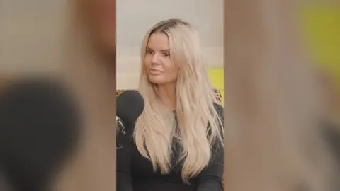 Kerry Katona has long blonde hair and is wearing a black top. She is sitting in front of a black microphone.