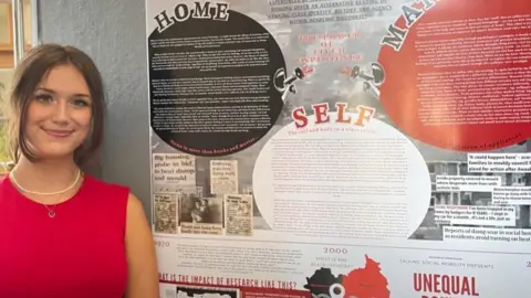 Mia Taylor-Seal A young woman with brown hair tied back and wearing a red top. She is standing next to a display she has created about working class people. Headings on the display read "self", "home" and "the power of lived experience"