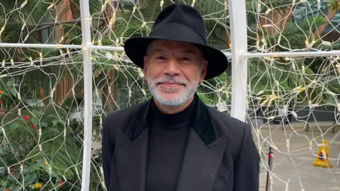 Tom MacDougall/BBC Russell McFarlane, a mixed-race man in his 60s with a short, white beard. He's smiling and wears an all-black suit with a black Panama hat.