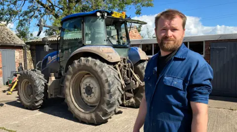 John Story is wearing blue overalls and standing in front of a blue tractor in a farmyard. He has short hair and a beard and moustache. Behind him are low level farm buildings with a tree behind them.