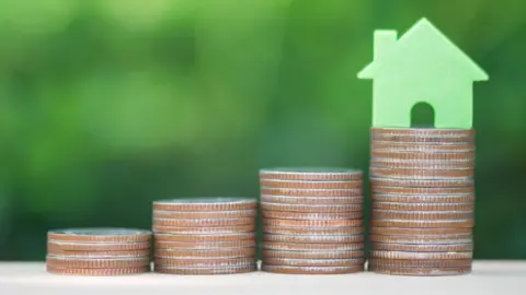 There are four stacks of coins getting taller as you move from left to right across the image. On the tallest stack of coins there is a little light green plastic house. 