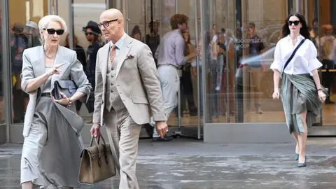 Getty Images On the left, Meryl Streep is seen wearing a light grey blazer and skirt with a brown belt and dark sunglasses. Her hair is in a quiff and Stanley Tucci is next to her in a beige suit as they walk away from Anne Hathaway's character in the background