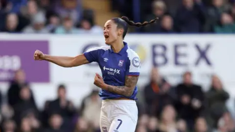 Getty Images Natasha Thomas is pictured in the midst of a football game. She is celebrating with her right arm punching out. She is shout while jumping in the air. Supporters can be seen in the background watching on.