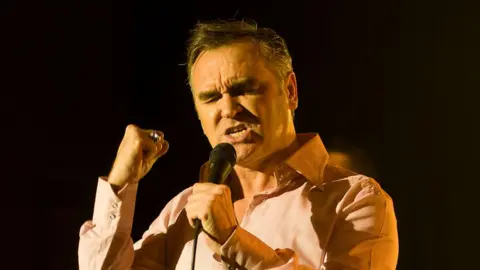 BBC Morrissey on stage holding a microphone, clenching his first with his eyes closed as he sings against a blacked out backdrop.