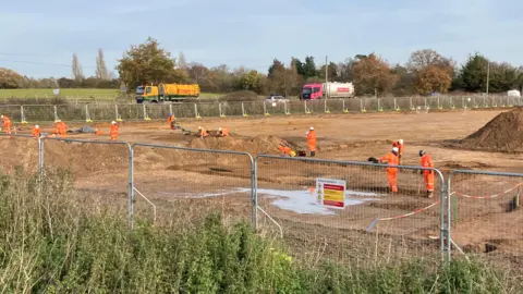 GUY CAMPBELL/BBC Archaeologists wearing hats and orange fluorescent jackets and trousers are working in an industrial landscape of bare sandy soil surrounded by a metal fence   