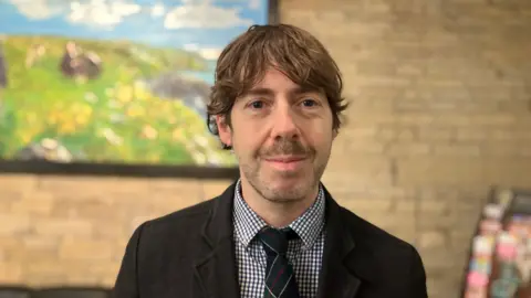 Elizabeth Baines/BBC A man with brown hair wears a suit. He stands in front of a york stone wall which is blurred