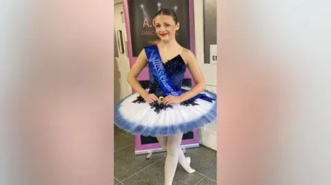Family photo A photo of Eleanor Hague. She has brown hair in a bun and is wearing a tutu and is smiling