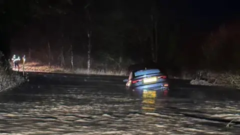 A flooded road with the image taken at night. A stranded car is submerged in the water and has been bathed in headlight from an approaching car. In the distance, a person can be seen looking into the water.