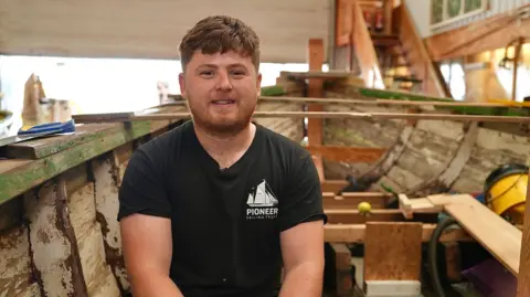 Martin Giles/BBC Noah has ginger hair and beard. He is wearing a black, Pioneer Sailing Trust-branded T-shirt, and is sitting inside a wooden boat that is undergoing restoration. 
