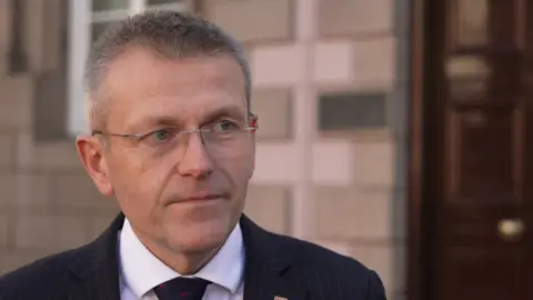BBC Philip Ozouf looks toward the camera. He is wearing a dark suit and tie with a white shirt. He has a lanyard around his neck. He has rimless glasses and very short grey hair.