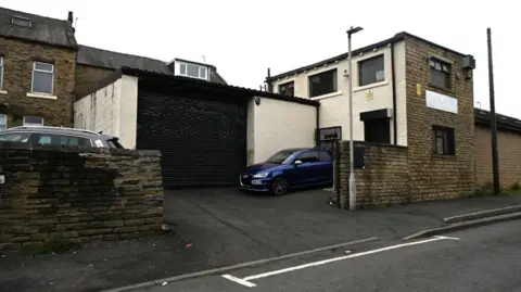 A garage which has a black roller shutter and a blue car parked on the drive outside. It is behind what looks like a row of stone terraced houses. 