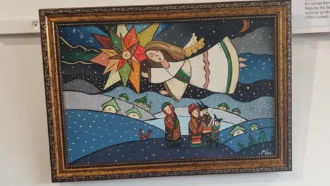 Michael Velychko A painting with a brown frame. In the painting there is an angel flying over homes and three men. There is a dog in the painting and a star.