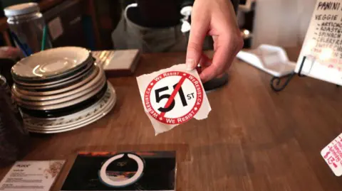 Getty Images A barista hands out a sticker against threatened 51st state at a coffee shop in Toronto, Canada on March 5, 2025. A coffee shop in Toronto renames its 'Americano' to 'Canadiano' and hands out stickers against threatened 51st state.