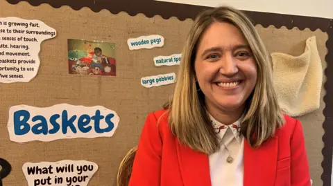 A woman with blond shoulder length hair is smiling. She is wearing a white blouse with a gold necklace and a red suit jacket. She is stood in front of a noticeboard which has words pinned on about babies and toddlers