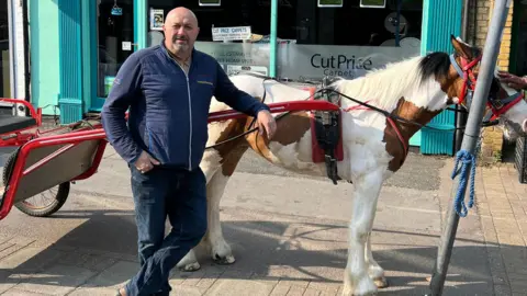 Man wears a blue jumper stands alongside a horse and cart outside a shop front. the shop sells carpets. The horse is white and brown. A hand is stroking the horse's forelock. The cart is an open one and is painted red. 