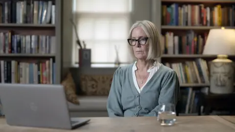 A photo of Dr Douglas reviewing the footage. She has grey hair in a bob-style and wears glasses. She has a grey cardigan and a white blouse with a necklace. She is in a room with lots of books in bookcases. There is a laptop on a desk that she is looking at. 
