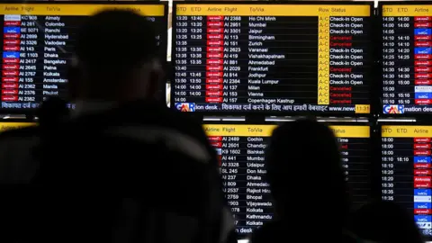 Passengers look at the screen displaying flight details as several flights are cancelled due to regional airspace closures following escalating tensions and reported military strikes involving Iran, Israel, and the United States in New Delhi, India, on March 1, 2026.