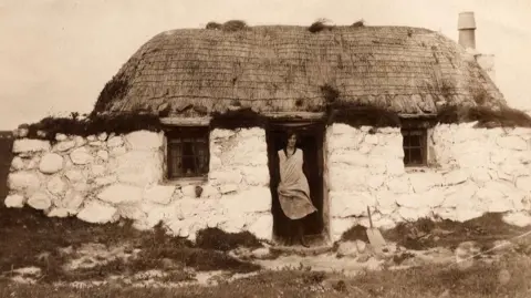 An Iodhlann A woman standing in the doorway of thatched roof cottage