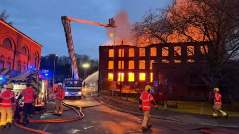 Orange flames can be seen inside the three-storey brick mill as a firefighter tacklles the blaze from an aerial platform at sunrise. Firefighters can also be seen on the road as the sun rises.