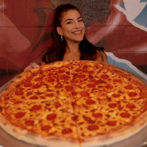 Leah Shutkever A woman sits down with an extra large pizza in front of her. She has straight dark hair and she is smiling.