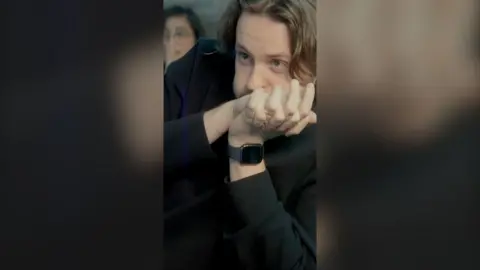 Man with long brown hair clasps hands against his face as he waits anxiously.