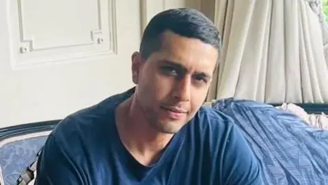 Kamran Aman wearing a blue top and sitting a sofa with a blue blanket behind him. He is looking at the camera and smiling. 