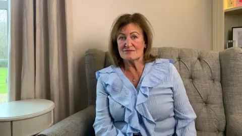 Marion Tyler is sitting on a grey armchair. She is looking directly at the camera with a sombre expression. She has brown shoulder length hair and red lipstick on. She is wearing a silver necklace. 