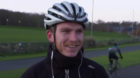 A man wearing a silver cycling helmet and a black waterproof jacket. He has a ginger moustache and mutton chops and has white headphones hanging out of his jacket. 