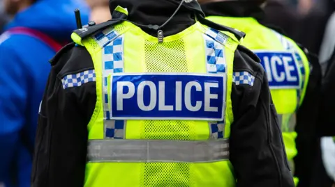 Two police officers seen from behind wearing high-visibility yellow jackets with blue and white chequered markings and 'POLICE' patches on the back. The officer in the foreground has a radio clipped to the shoulder of a black fleece underneath, with a coiled earpiece wire visible. The image is taken in a crowd setting with other figures partially visible in the background.