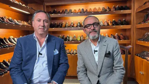 Dean Girling (left) and Tony Gaziano (right) sit side-by-side in a room surrounded by shelves of bespoke shoes. Mr Girling is in a blue suit and shirt with a light blue pocket square. Mr Gaziano is in a black and white chequered jacket with a black pocket square. He is also wearing black framed glasses.