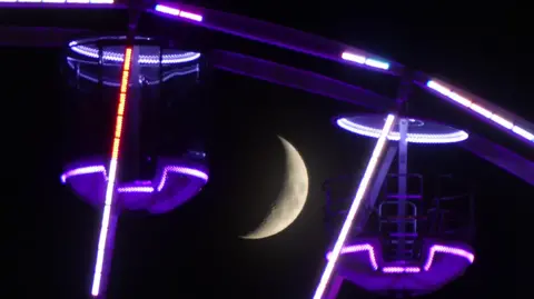 David Pritchard The moon in between two cars on a big wheel in Glasgow. The moon is in the centre of the image. The two cars on either side are glowing purple neon.