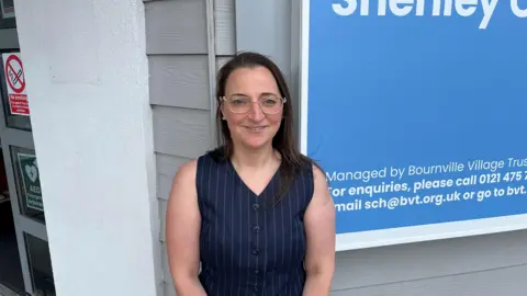 A woman with brown hair and clear glasses smiles at the camera in front of a community building. She wears a blue pinstriped waistcoat with a v-neck.