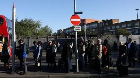 Passengers queue in a line at a bus stop with a red bus just in shot going in the opposite direction. There is a block of flats in the background.
Some of the travellers have suitcases with them.