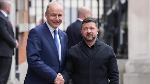 Taosieach Micheál Martin and President Volodymyr Zelensky. They are shaking hands. Martin is wearing a navy suit and Zelensky is wearing a black jacket. Martin is smiling. 