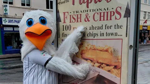Visit Weston-super-Mare Mascot Sammy the Seagull, who wears a white and black striped Weston-super-Mare AFC shirt, poses with a large sign for "traditional fish and chips".