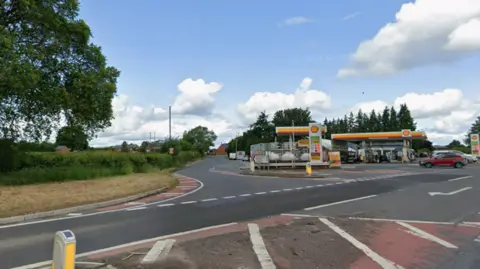 Google Maps A left hand turn on a main road with a petrol station also on the right of the turn. The sky is blue and slightly cloudy. 