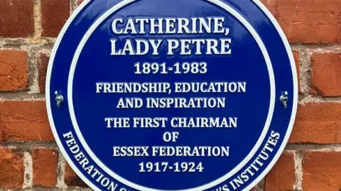 A blue plaque fixed to a red-brick wall. It reads CATHERINE LADY PETRE. 1891-1983. FRIENDSHIP, EDUCATION AND INSPIRATION. THE FIRST CHAIRMAN OF ESSEX FEDERATION 1917-1924.