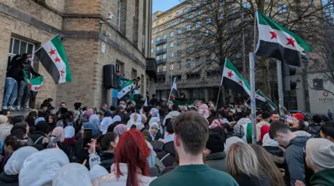 A wide shot of a large crowd of people gathered outside the BBC in Belfast celebrating the fall of the Assad regime. There are about seven flags of the new Syrian regime visible - green, white and black horizontal stripes with three red stars in the middle of the white stripe.