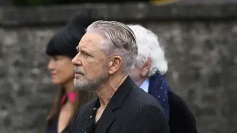 PA Media Adam Clayton, with grey hair and grey beard, wearing a dark suit and dark shirt, at the funeral of Lord Mount Charles.